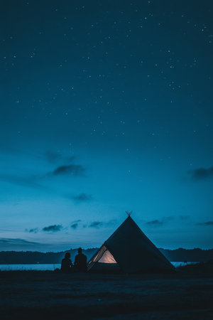 Silhouette of Illuminated Tent By Lake At Dusk Under Deep Blue Night Sky Full of Starsの素材