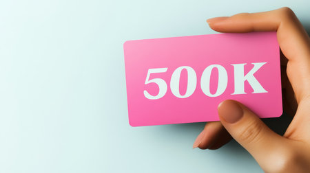Woman s Hand Holding Pink Card Displaying 500K Text, Celebrating Success on a Pastel Backgroundの素材