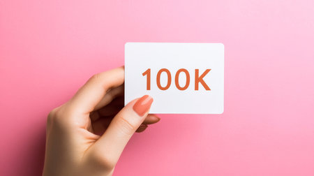 Woman s hand holding a white card with 100K in rust-orange numerals on a pastel pink backgroundの素材