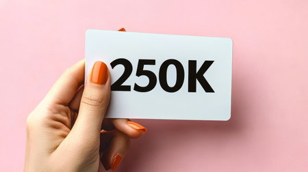 Woman s Hand with Orange Nails Holds Card Displaying 250K on Pastel Pink Backgroundの素材