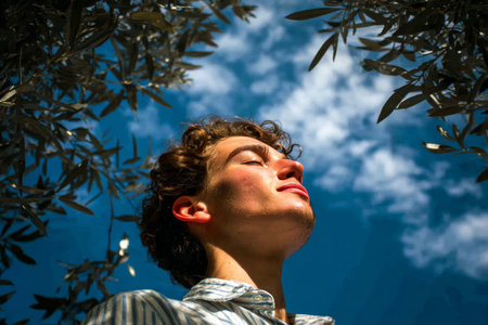 Relaxing young man enjoys the warmth of the sun in a vibrant green forest under a clear blue sky.の素材