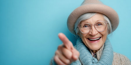 Joyful senior female in a stylish hat and eyewear, playfully posing against a pastel backdrop.の素材