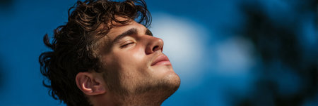 Young man enjoying a peaceful moment outdoors, absorbing sunlight and gazing at the clear blue sky.の素材