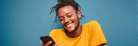 Attractive young man with cool dreadlocks and cheerful smile using mobile phone against blue wallの素材