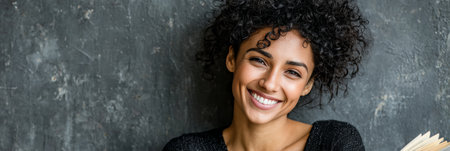 Beautiful woman with curly hair reads a book in a studio, smiling at the camera, looking relaxed.の素材