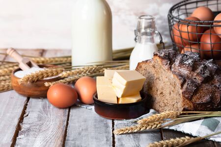 Concept of homemade bread, natural farm products, domestic production. Healthy and tasty organic food. Eggs, butter, bread, milk, flour. Wheat spica as decor. Closeup, wooden backgroundの写真素材