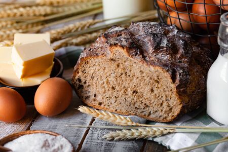 Concept of homemade bread, natural farm products, domestic production. Healthy and tasty organic food. Eggs, butter, bread, milk, flour. Wheat spica as decor. Closeup, wooden backgroundの写真素材