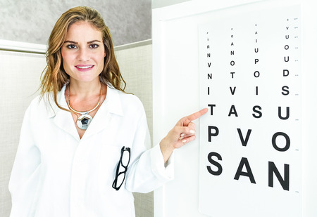 Female oculist doctor pointing at eye sight test chart and looking at camera - Optic medical concept in private clinic studio with young woman welcoming and cheering at patientの写真素材