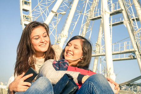 Best friends enjoying time together - Young people with smartphone music playlist in autumn sunny day - New trends and technology concept with hipster girlfriends having fun outdoors at ferris wheelの写真素材