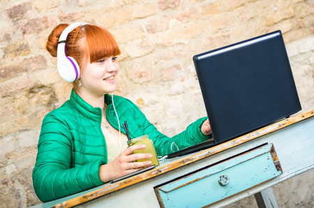 Hipster redhead woman with headphone using laptop drinking cocktail and having fun at work - Modern concept of professional engagement connected with happy and productive attitude - Focus on girl faceの写真素材