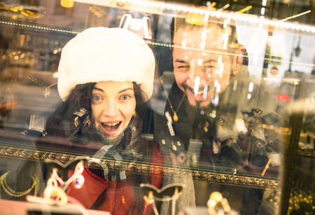 Modern hipster couple shopping on winter cloth pointing jewelry store window display - Consumerism concept with young fashion people looking for Christmas present at urban city tour - Focus on womanの写真素材
