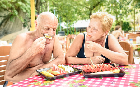 Senior couple having fun eating seasonal fruit in thai restaurant bar outdoors - Mature man and woman on active elderly vacation - Happy retirement concept with people together - Warm shadow filterの写真素材