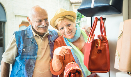 Senior couple shopping at fashion bag store with wife pointing showcase to husband - Active elderly concept with mature man and woman having fun in city - Happy retired people moments on vivid colorsの写真素材