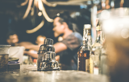 Blurred defocused side view of barman and people drinking and having fun at cocktail bar - Social gathering concept with people enjoying time together - Warm retro contrast filter with focus on shaker