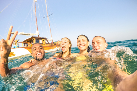 Young multiracial friends taking selfie and swimming on sailing boat sea trip - Rich happy guys and girls having fun in summer party day - Exclusive vacation concept - Bright afternoon  warm filterの写真素材