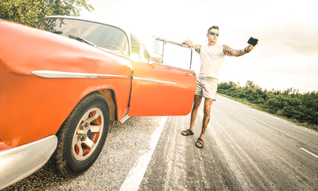 Young hipster fashion man with tattoo taking selfie with vintage car during road trip in Cuba - Travel wanderlust concept always connected on social media influencer lifestyle - Bright retro filterの写真素材