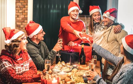 Friends group wearing santa hat giving each other Christmas presents - Champagne wine toast at home x mas dinner - Holiday concept with young people sharing time together and having fun on winter timeの写真素材