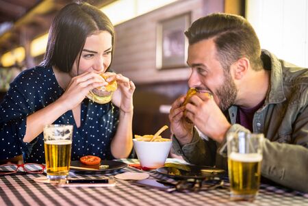 Happy couple having fun eating burger at restaurant pub fast food - Young people enjoying moment at indoor diner location - Relationship concept with focus on girl face with warm vivid filterの写真素材