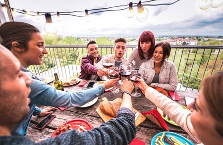 Wide angle view of happy people toasting red wine together at rooftop party in open air villa - Young friends eating bar-b-q food at restaurant patio - Cool friendship concept on warm vivid filterの写真素材