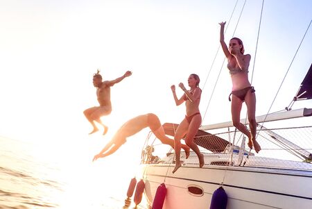 Young millenial friends jumping from sailboat at sea ocean trip - Guys and girls having summer fun together at sail boat party day - Luxury excursion concept on bright vivid filter with soft focusの写真素材