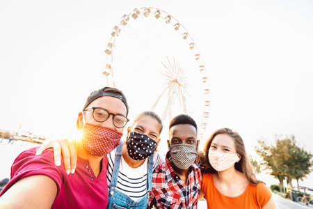 Multiracial milenial students taking selfie protected by face masks - New normal travel concept with young people having safe fun together at ferris wheel - Bright sunshine filter with tilted angleの写真素材