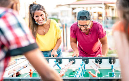 Multiracial friends play kicker table football at open space bar - New normal lifestyle concept with happy milenials having fun together with open face mask - Bright vivid filter with focus on guyの写真素材