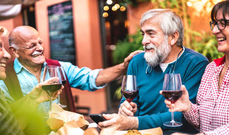 Happy senior friends having fun drinking red wine at dinner party - Retired people eating at restaurant together - Dinning lifestyle concept on warm vivid filter - Focus on bearded hipster manの写真素材