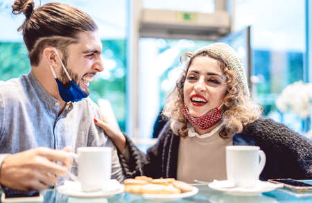 Happy couple wearing face mask having fun together at bar cafeteria - New normal lifestyle concept with young people on positive mood drinking american coffee - Bright azure filter with focus on womanの写真素材