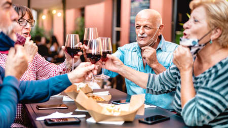 Senior friends toasting wine at restaurant bar wearing opened face mask - New normal lifestyle concept with happy mature people having fun together at garden party - Warm filter with focus on bald manの写真素材
