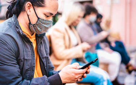 Asian guy using smart phone covered by face mask on Covid second wave - New normal lifestyle concept with milenial people watching news on mobile smartphone - Shallow depth of field with focus on eyeの写真素材