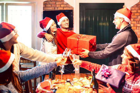 Friends with santa hat giving each other Christmas present - Champagne wine toast at house x mas dinner party - Holiday concept with people having fun together on winter time - Focus on central guyの写真素材