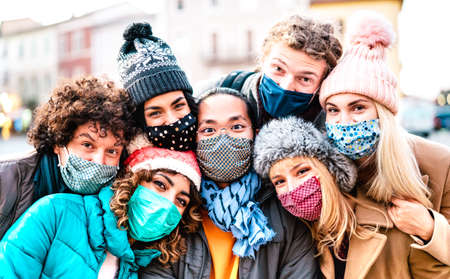 Multiracial friends taking selfie wearing face mask and winter clothes - New normal lifestyle concept with young people having fun together outside - Bright filter with focus on central asian guyの写真素材