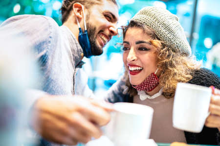 Happy couple with face mask having fun together at bar cafeteria - New normal lifestyle concept with young people talking and drinking american coffee - Bright azure filter with focus on woman eyeの写真素材