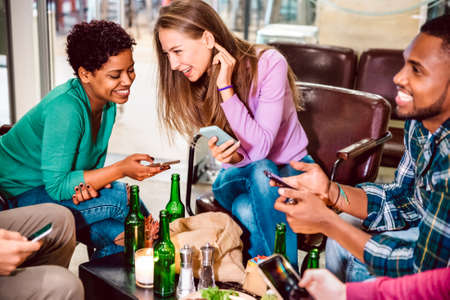 Multiracial friends drinking beer and having fun with mobile smart phones at cocktail bar restaurant - Friendship concept with milenial people enjoying time together - Focus on central caucasian womanの写真素材