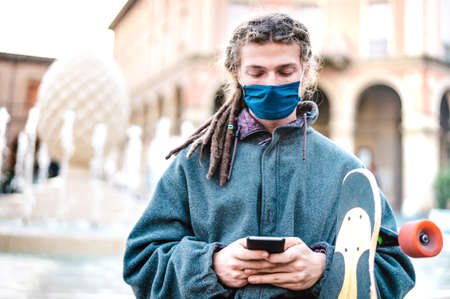 Unhappy guy with protective mask using tracking app on mobile smartphone - Young worried millenial sharing content on social media - New normal lifestyle concept - Bright filter with focus on faceの写真素材