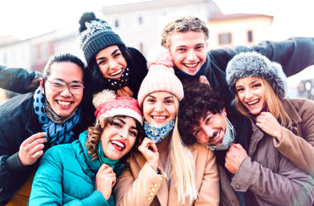 Multiracial friends taking selfie with open face mask and winter clothes - New normal friendship concept with milenial people having fun together outside - Bright filter with focus on central womanの写真素材