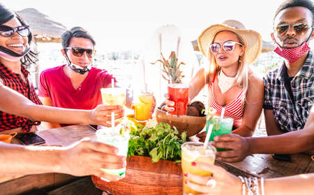 Young multicultural people toasting at beach cocktail bar with open face mask - New normal summer concept with milenials having fun together cheering drinks - Vivid filter with focus on girl with hatの写真素材