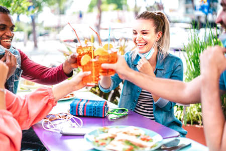 Young people drinking at cocktail bar wearing face masks - New normal friendship concept with friends having fun together toasting drinks at restaurant - Bright filter with focus on right woman