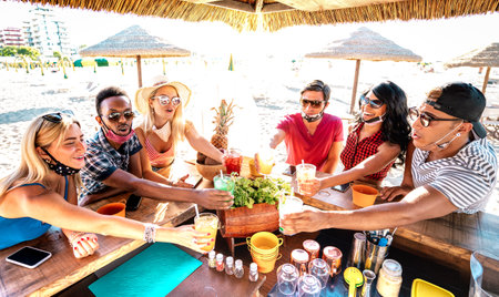 Young trendy people toasting at beach cocktail bar chiringuito with open mask - New normal summer life style concept with friends having fun together cheering drinks - Warm bright backlight filterの写真素材