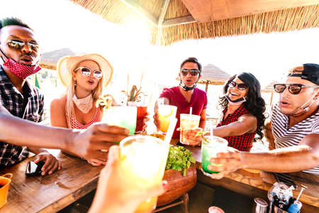 Young multiracial people toasting at beach cocktail bar with open face mask - New normal life style summer concept with happy friends having fun together cheering drinks - Warm backlight filterの写真素材