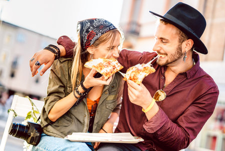 Young hipster couple eating pizza at bar restaurant outdoors - Happy relationship concept with millenial boyfriend and girlfriend having fun moments together - Bright vivid filter with focus on facesの写真素材