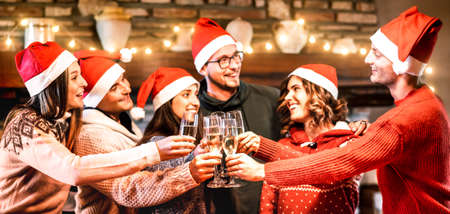 Young friends with santa hats celebrating Christmas with champagne wine toast at home reunion - Winter holidays concept with happy millenials enjoying time and having fun together - Focus on glassesの写真素材