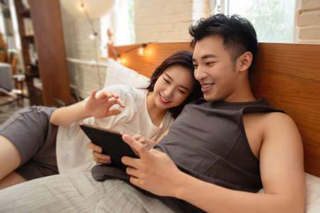 Happy young Asian couple using mobile phone while lying on bed at homeの写真素材