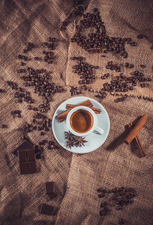 dark way of white cup of coffee with milk and aromatic spices: cinnamon sticks, star anise, dried slices of lemon and coffee beans on a napkin of burlapの写真素材