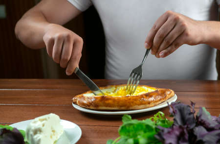Man eating adjar Khachapuri. Georgian national pie khachapuri with egg and cheese in the white plate...
