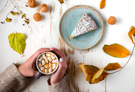 Top view of female hands holding marshmallows on shabby table with piece of cake, walnuts and scattered leavesの写真素材