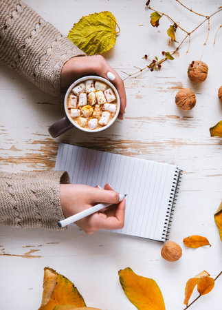 Top view of female hands holding cup of cocoa with marshmallow on shabby table with yellow scattered...