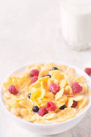bowl of corn granola with milk, fresh raspberries, blueberries. milk in a jug and a glass on white wooden board for healthy breakfastの写真素材