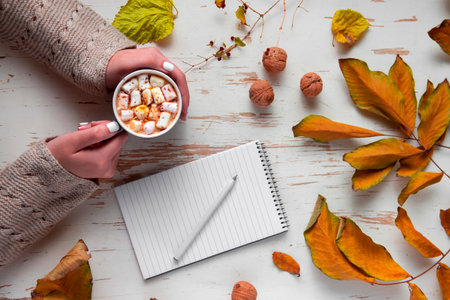 Top view of female hands holding cup of cocoa with marshmallow on shabby table with yellow scattered...
