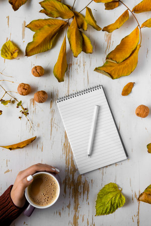 Top view of female hands holding cup of coffee on shabby table with yellow scattered leaves, walnuts...